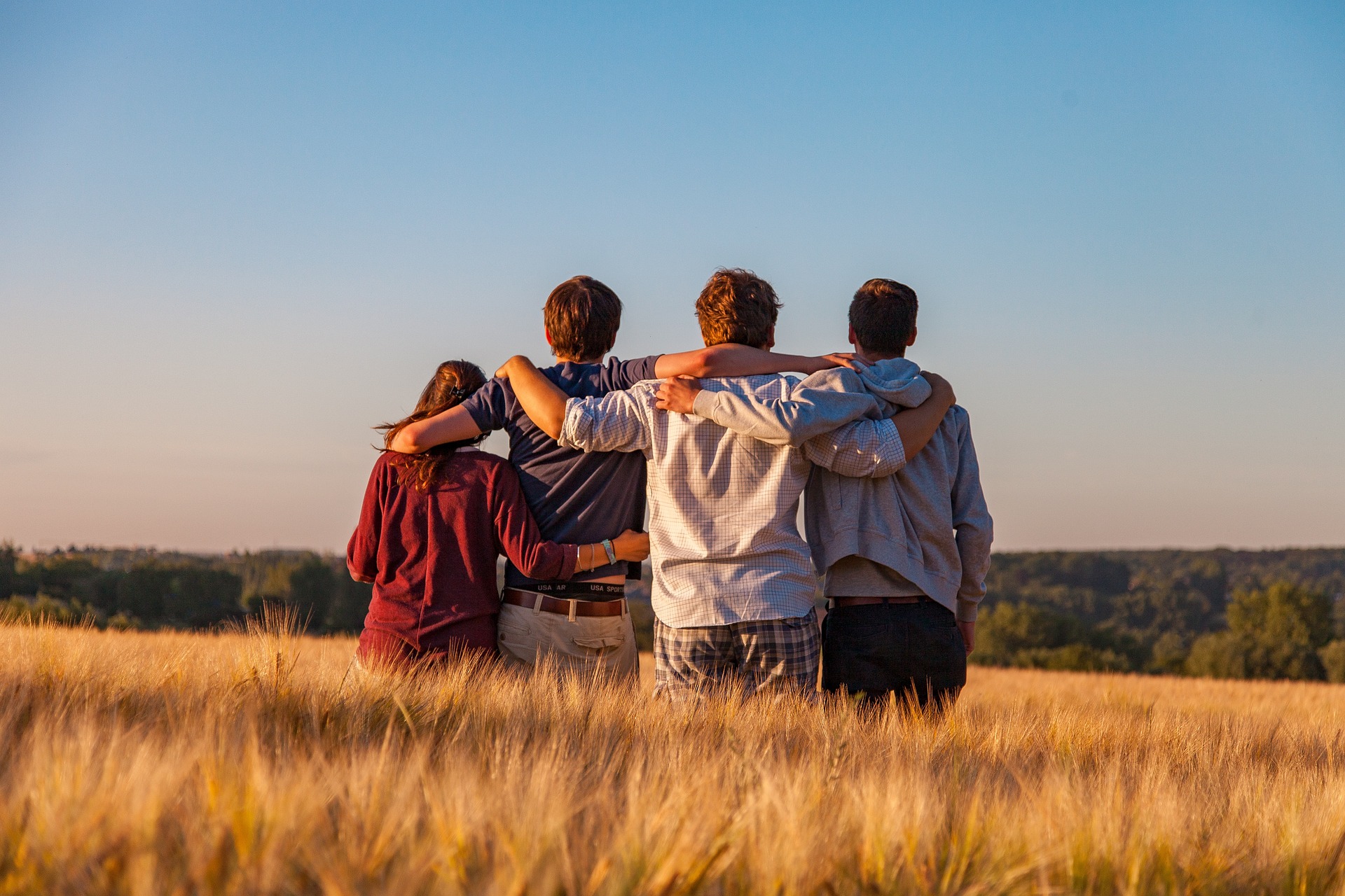 Four young adults standing together with arms around each other in a golden field at sunset